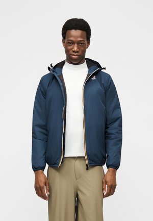 Bărbat purtând o jachetă bleumarin cu glugă peste un pulover alb cu guler înalt și pantaloni bej, stând în picioare pe un fundal simplu gri deschis.