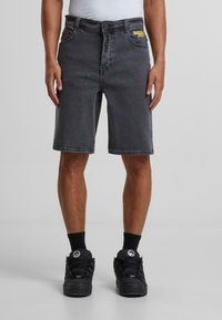 Shorts en denim gris à coupe droite, dotés d'un design classique à cinq poches et d'un patch logo subtil près de la taille. Portés avec des baskets noires.