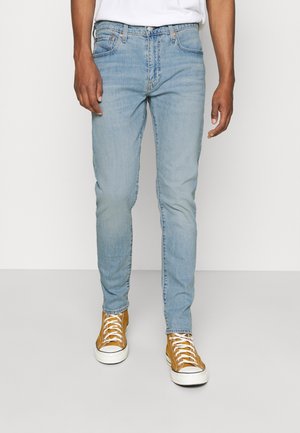 Mand iført lyseblå slim-fit jeans, hvid T-shirt og brune høje sneakers står foran en ensfarvet baggrund.