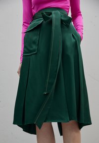 Žena v sukni s vysokým pasem v tmavě zelené barvě se zavazováním vpředu a velkou kapsou, kterou doplňuje svetrem s dlouhým rukávem v jasně růžové barvě.