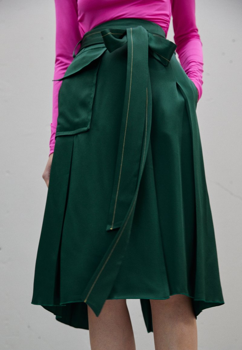 Žena v sukni s vysokým pasem v tmavě zelené barvě se zavazováním vpředu a velkou kapsou, kterou doplňuje svetrem s dlouhým rukávem v jasně růžové barvě.