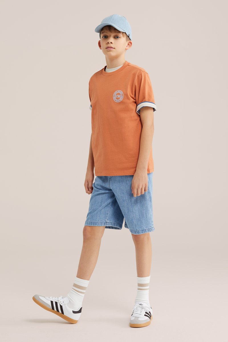 Garçon portant une casquette bleu clair, un t-shirt orange, un short en jean, des chaussettes blanches rayées et des baskets blanches, faisant un pas en avant sur un fond uni.