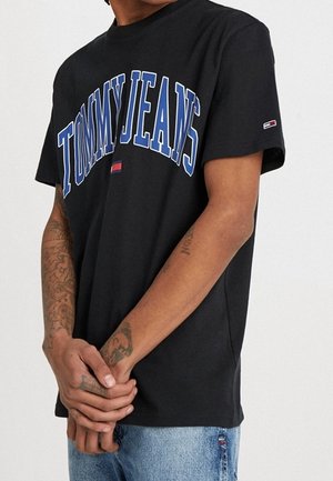 Zwart katoenen T-shirt met "TOMMY JEANS" in grote blauwe letters. Korte mouwen, ronde hals en logolabel op de linkermouw.