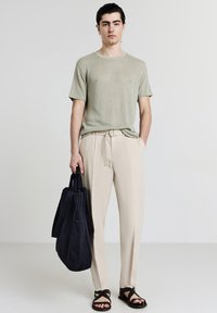 Giovane uomo in piedi su uno sfondo neutro, indossa una maglietta corta verde chiaro, pantaloni beige e sandali neri, tiene in mano una grande borsa a tracolla nera.