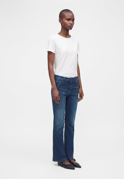 Femme aux cheveux courts portant une chemise blanche à manches courtes, un jean bleu droit et des ballerines noires pointues, debout devant un fond blanc.