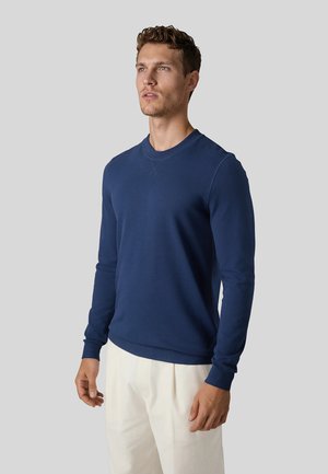 Ung mand med kort krøllet hår iført en marineblå langærmet sweater og hvide bukser, stående foran en ensfarvet grå baggrund.