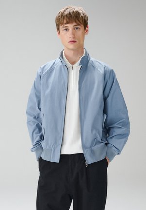 Ung mand iført en lyseblå lynlåsjakke over en hvid lynlås sweater med krave og sorte bukser, stående med hænderne i lommerne mod grå baggrund.