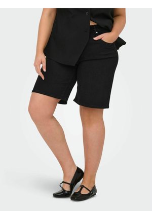 Person, die schwarze knielange Shorts mit vorderen Taschen, ein ärmelloses schwarzes Oberteil und schwarze Mary Jane Ballerinas trägt und mit einer Hand in der Tasche steht.