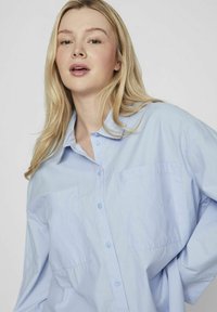 Camisa de botones azul claro hecha de una tela suave, con un cuello clásico y dos bolsillos en el pecho. Corte holgado con mangas largas.