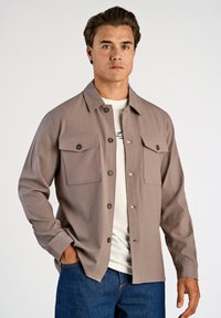 Brun skjorta med krage och två bröstfickor, knäppning fram och långa ärmar, i kombination med en vit t-shirt, bärs över ett par blå jeans.