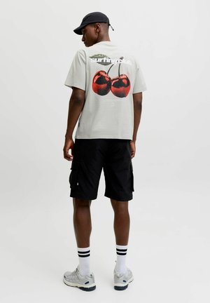 Mand iført en lysegrå T-shirt med stor rød kirsebær-grafik og tekst på ryggen, sorte shorts, hvide sokker med striber, sneakers og en sort kasket.
