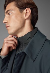 Jachetă închisă la culoare, cu un guler înalt și fermoar vizibil. Textură netedă, mată, accentuată de o strat interior contrastant. Vedere de profil în prim-plan.