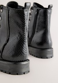 Mustat nilkkurit, joissa on kuvioitu matelija-aiheinen pinta, sivuvetoketju ja nauhalliset kiinnitykset. Paksu kumipohja, jossa on kulutuspintadetalji.