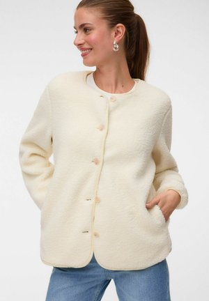 Femme portant une veste polaire crème boutonnée avec des poches, un jean bleu et des boucles d’oreilles argentées en forme d’anneau, souriante et regardant sur le côté.
