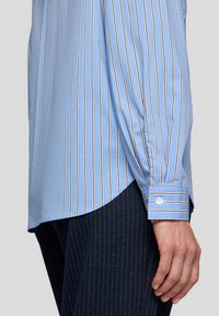 Light blue striped shirt with thin white and dark stripes, long sleeves, button cuffs, and a curved hem. Fabric appears smooth and textured.