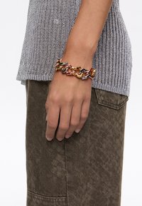 Pulsera de cadena metálica multicolor que presenta eslabones en oro, plata y bronce, llevada en una mano sobre una blusa texturizada gris y pantalones marrones.