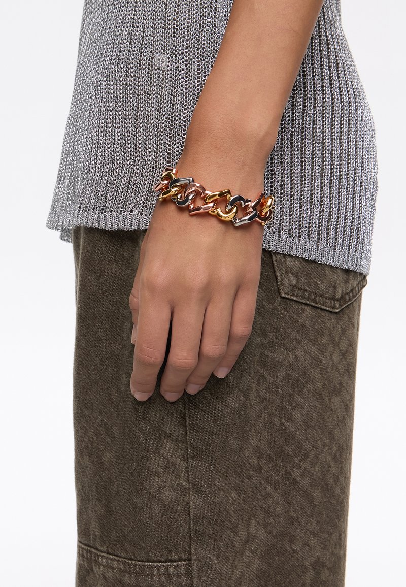 Pulsera de cadena metálica multicolor que presenta eslabones en oro, plata y bronce, llevada en una mano sobre una blusa texturizada gris y pantalones marrones.