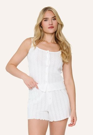 Blonde Frau trägt weißen ärmellosen, geknöpften Stricktop und passende Strickshorts, posiert mit einer Hand auf der Hüfte.