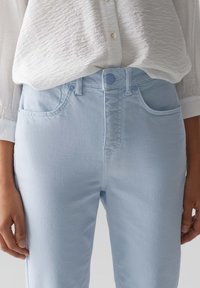 Jean en denim bleu clair à taille haute, avec passants pour ceinture et poches avant. Le tissu a une texture lisse et des détails de couture soignés.