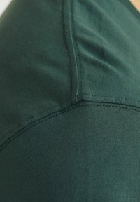 Tessuto di maglietta verde scuro che mostra un primo piano della cucitura della spalla, con una texture liscia e dettagli di cucitura a contrasto.