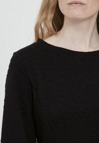 Mustat pitkät hihat -paita, jossa on teksturoitu, kuvioitu kangas. Pyöreä kaula-aukko ja tasainen väri, joka esittelee hienovaraista kohokuviointia.