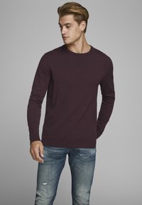 Suéter burdeos de cuello redondo hecho de una tela suave, con mangas largas y puños acanalados, combinado con jeans azules desgastados.