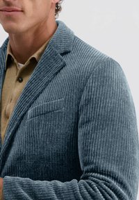 Gelaagde blauwe corduroy blazer met een geknipte kraag, een enkel borstzakje en een getailleerd silhouet, gedragen over een tan kleurig shirt.