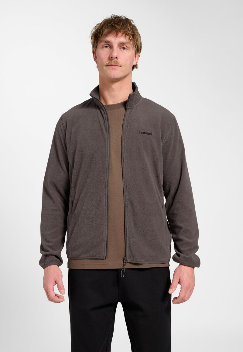 Mann trägt dunkelgraue Fleecejacke mit Reißverschluss über braunem Hemd und schwarzer Hose, steht vor einem schlichten hellen Hintergrund.