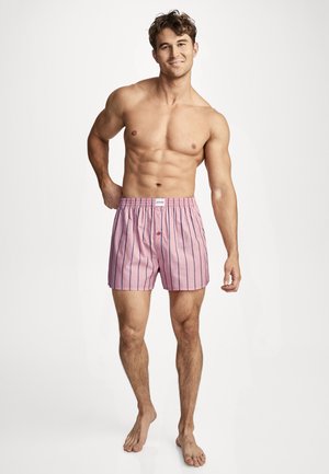 Muscular man standing barefoot wearing pink striped boxer shorts, smiling, with one hand behind back and one hand relaxed by side.
