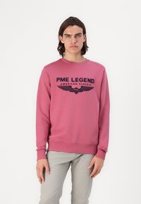 Różowa bluza z okrągłym dekoltem, z napisem "PME LEGEND AMERICAN CLASSIC" i grafiką skrzydeł. Długie rękawy, ściągacze przy rękawach i u dołu.