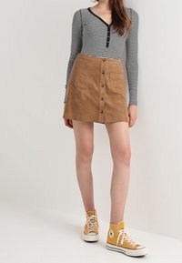 Bruine suède mini-jurk met knopen aan de voorkant en zakken, gecombineerd met een zwart-wit gestreepte blouse met lange mouwen en gele hoge sneakers.