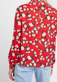 Blouse rouge avec motif floral présentant des roses blanches et des accents bleus, encolure froncée, manches longues et fermeture à boutons dans le dos.