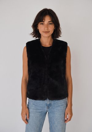Donna con capelli corti e castani che indossa un gilet nero senza maniche in pelliccia sintetica e jeans azzurri, in piedi contro uno sfondo chiaro e uniforme.