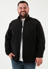 Chaqueta de forro polar negra con cierre delantero, dos bolsillos laterales y un bolsillo en el pecho. Cuenta con un collar alzado y una textura suave.