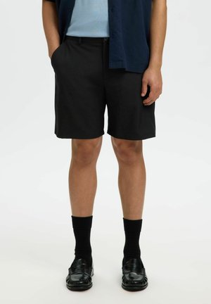 Homme portant un short noir sur mesure, des chaussettes noires et des mocassins en cuir noir, debout avec une main dans la poche devant un fond uni.