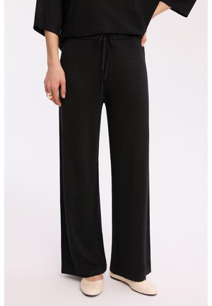 Czarne spodnie o szerokich nogawkach z wiązaniem w talii, noszone z beżowymi płaskimi butami i czarnym topem, skupienie na dolnej części tułowia i nogach.