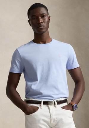 Mand iført lyseblå kortærmet skjorte, hvide bukser, sort bælte og brun urrem, stående med hænderne i lommerne mod en neutral baggrund.