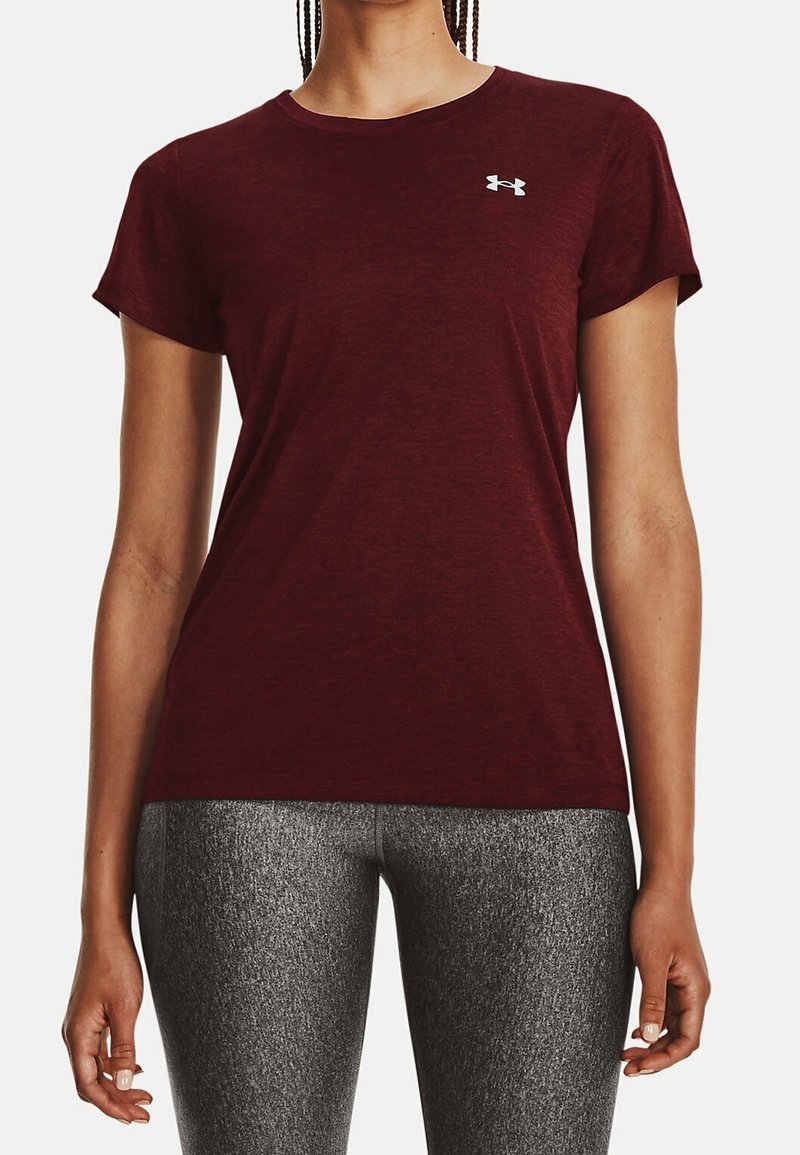 Kobieta w bordo koszulce Under Armour z krótkim rękawem i szarych teksturowanych legginsach sportowych, stojąca z rękami swobodnie opuszczonymi wzdłuż ciała.