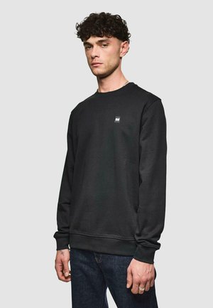 Black sweatshirt with long sleeves, round neckline, and visible stitching. Features a small, square logo on the chest. Worn over dark jeans.