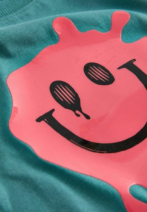 Türkisfarbenes T-Shirt mit einem glänzenden pinken Spritzdesign, versehen mit einem schwarzen Smiley-Gesicht und gestreiften Augen. Weiche Baumwolltextur, lässiger Stil.