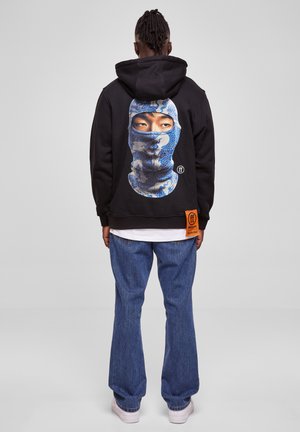 Svart hettegenser med et stort grafisk motiv av et maskert ansikt i blått og hvitt. Har et oransje merke på siden. Bæres med blå jeans.
