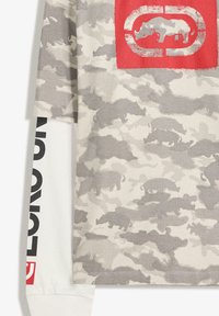 T-shirt grigio con motivo camouflage, caratterizzato da un logo a scatola rossa con la silhouette di un rinoceronte e maniche lunghe bianche con accenti di testo neri.