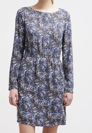 Kobieta w sukience z długim rękawem, sięgającej powyżej kolana, z abstrakcyjnym wzorem w kolorach niebieskim, czarnym i białym oraz lekko marszczoną talią.