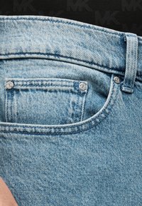 Μπλε τζιν παντελόνι με μπροστινή τσέπη, εμφανείς ραφές και μεταλλικά καρφιά. Το ύφασμα έχει ξεθωριασμένη, υφασμένη όψη.