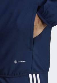 Donkerblauwe jas met een gladde textuur, voorzien van een zijzak en een wit "AEROREADY" logo. Het ontwerp omvat drie witte strepen nabij de zoom.