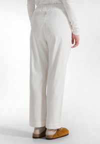 Witte geribde broek met een elastische tailleband, wijde pijpen en zichtbare stiksels. Gemaakt in combinatie met kastanjebruine instappers en beige sokken.