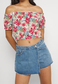Donna che indossa un crop top bianco a spalle scoperte con stampa floreale rossa e una gonna mini asimmetrica in denim blu a vita alta.
