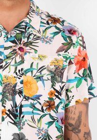 Chemise à manches courtes en tissu léger arborant un imprimé floral vif. Principalement rose pâle avec des accents orange, violet et vert.