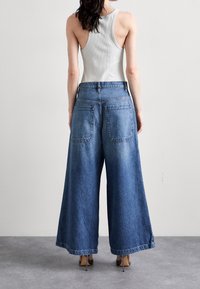 Jeans de denim de pierna ancha en azul claro, con una cintura alta, dos bolsillos traseros y un acabado suave; combinados con una camiseta de tirantes gris.