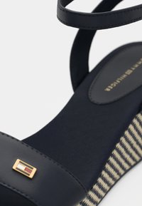 Schwarze Keilsandalen mit glattem Obermaterial, verstellbarem Knöchelriemen und Korksohle im Hahnentritt-Muster. Goldenes Logo-Detail am Riemen.